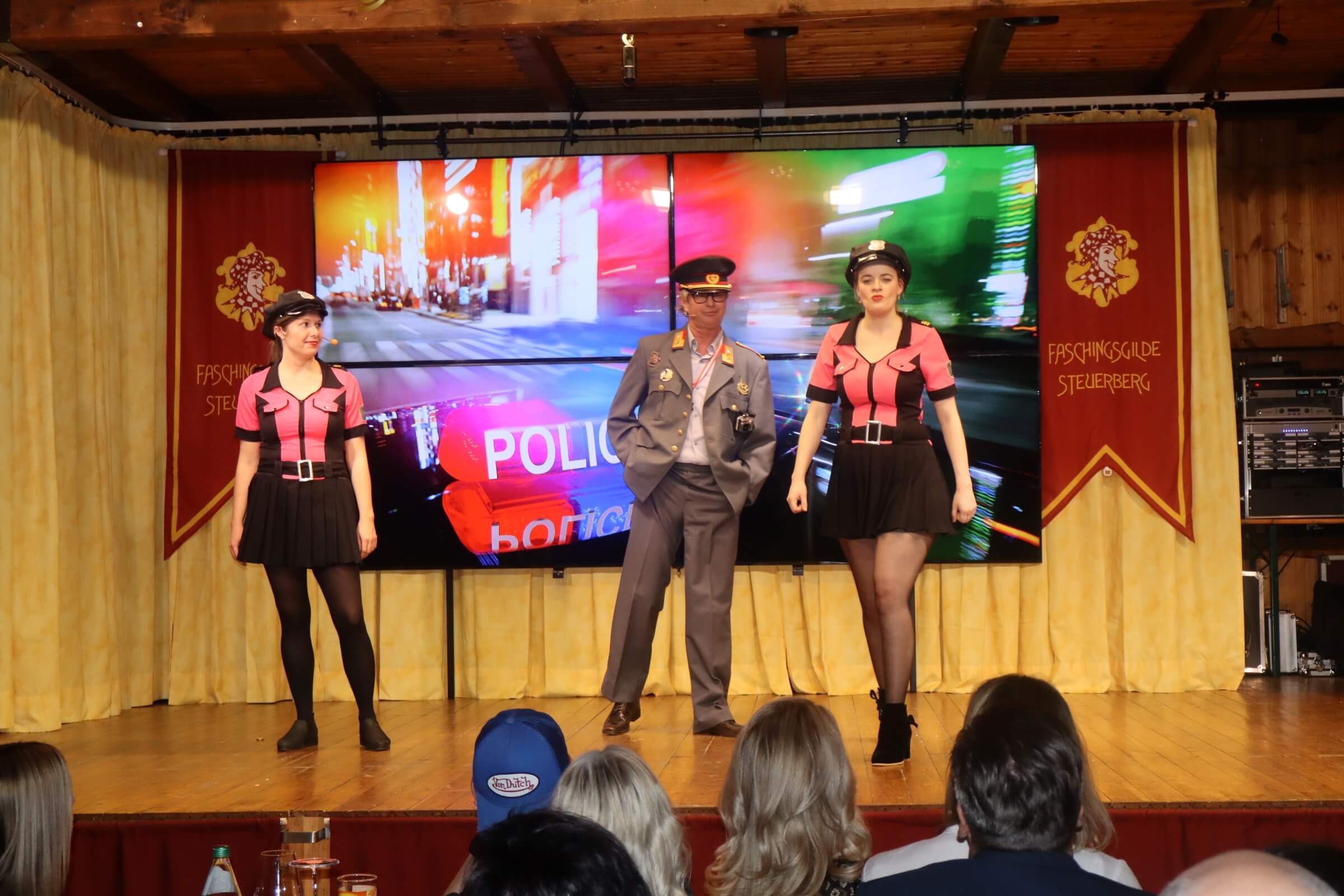 Humorvolle Bühnenszene mit drei Darstellern vor einer Stadtkulisse